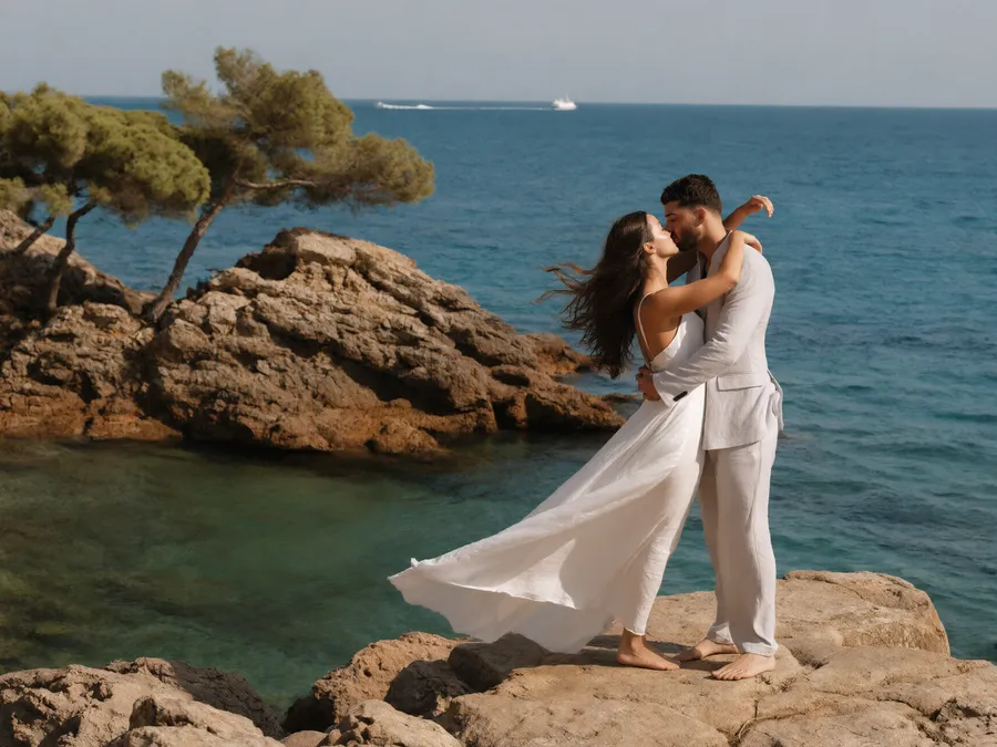 A newly married couple kissing barefoot on the rocks above a turquoise cove with pine trees